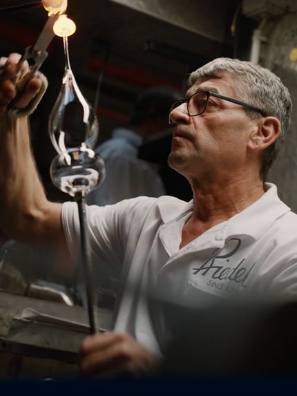 A master glassblower in a RIEDEL Manufaktur shirt shapes molten glass with precision tools, capturing the artistry and tradition of handmade glassmaking.