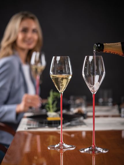 Two RIEDEL High Performance Champagne Red Stemmed glasses on a tabletop with Champagne filled in the left glass and Champagne pouring into the right glass. Behind is a lady holding a filled RIEDEL High Performance Champagne Red Stemmed glass.