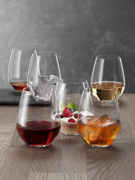 Six SPIEGELAU Authentis Casual tumblers on a wooden table, each of them filled differently. With red wine, with a soft drink on ice, with a creamy desssert, with white wine, with water and ice cubes and with a brown liquor.<br/>