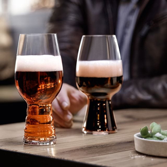 I bicchieri da birra artigianale SPIEGELAU riempiti di birra IPA e il bicchiere da Stout riempito di birra Stout in piedi su un tavolo di legno. Dietro di loro una mano che prende il bicchiere da Stout.<br/>