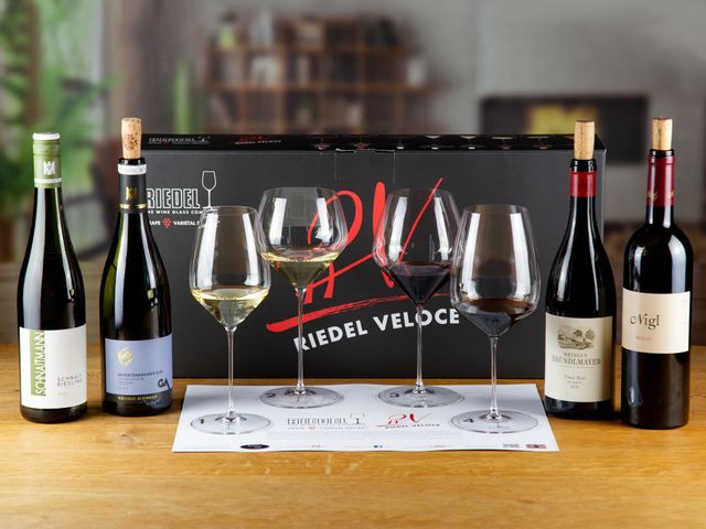 Tasting mat with 2 filled different white wine glasses and two filled different red wine glasses of the RIEDEL Veloce series in the background 2 different white wine bottles, two different red wine bottles and the packaging of the RIEDEL Veloce tasting set