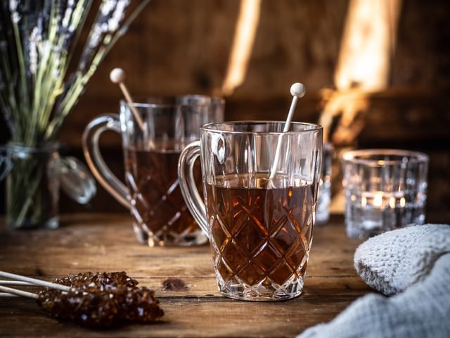 The NACHTMANN Noblesse tea mug with hot tea in it and rock candy. On the wooden table, there are more sticks with brown rock candy, a jar with lavender flowers, a second filled crystal tea mug and a shining tea light.<br/>