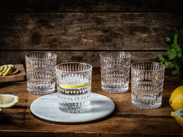 Four NACHTMANN Ethno tumblers on a wooden sideboard. One of them is filled with water and a slice of lemon and stands on a round stone platter. Around the tumblers are lemons, slices of lemon, and mint sprigs.<br/>
