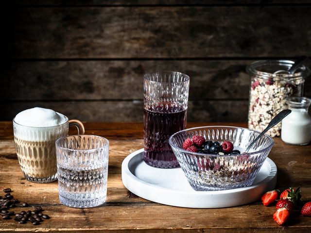 The NACHTMANN Ethno series on a wooden table. The hot beverage mug is filled with cappuccino, the tumbler with mineral water. The longdrink glass, filled with berry juice and the bowl, filled with a crunchy muesly, are standing on a white serving tray. In the background a jar with cereals and milk.<br/>
