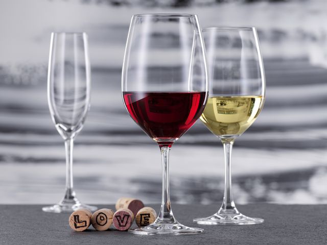 Un verre à bordeaux rempli de vin rouge et un verre à vin blanc rempli de vin blanc de la collection Winelovers de SPIEGELAU. À l'arrière-plan se trouve une flûte à champagne vide. Cinq bouchons de liège se trouvent derrière le verre de Bordeaux, quatre d'entre eux représentant les lettres du mot LOVE.<br/>