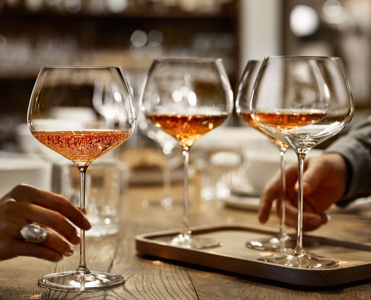 Eine Hand hält das SPIEGELAU Burgunderglas aus der Serie Willsberger Anniversary, gefüllt mit roséfarbenem Sekt. Weiter hinten auf dem Holztisch stehen weitere Gläser.<br/>