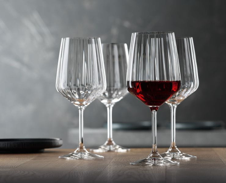 Four SPIEGELAU Lifestyle red wine glasses on a wooden table. One of them is filled with red wine.<br/>